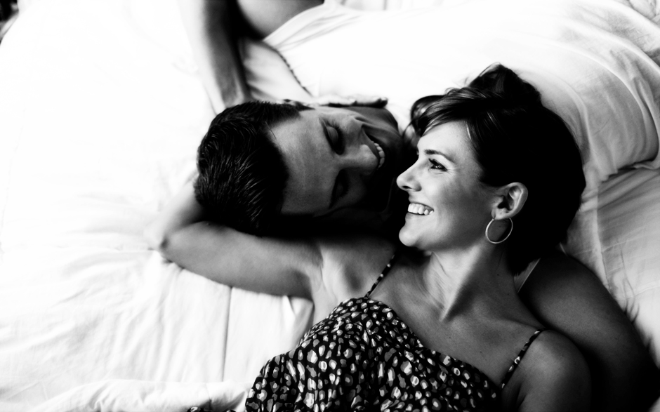 A couple smiles in bed at their Philadelphia apartment for engagement photos, photographed by Peter Van Beever