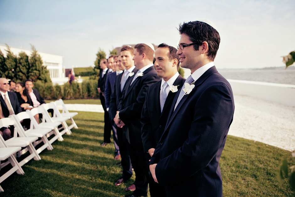 Groomsman stand in a line at the Belle Mer wedding in Newport Rhode Island, with the ocean and beach at their backs, wearing blue suits with white orchids