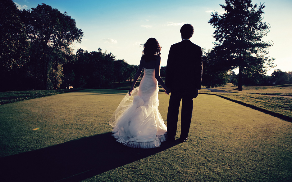 The bride and groom stand on the golf course in the setting sun on their wedding day, looking elegant