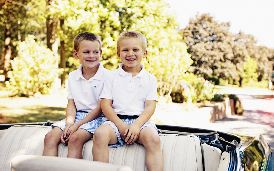 Two little boys wearing white polos and striped shorts sit in the back of a car with the roof down, on a sunny summer day